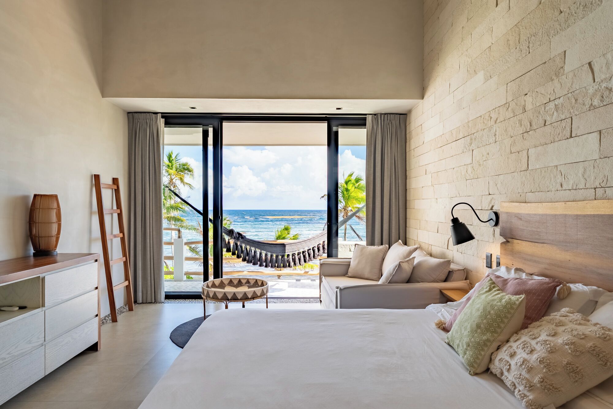 A bedroom with a large bed, a view of the ocean, a hammock, and a brick wall.