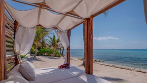 Una cabana sulla spiaggia con struttura in legno e tettoia in tessuto bianco, affacciata su una spiaggia sabbiosa e un mare cristallino.