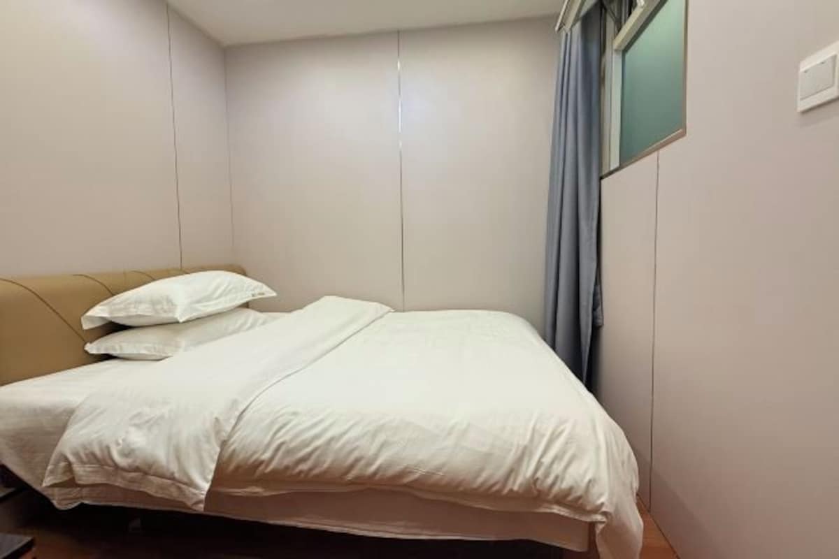 A single bed with a white bedspread and pillows, a wooden headboard, and a window with a curtain.