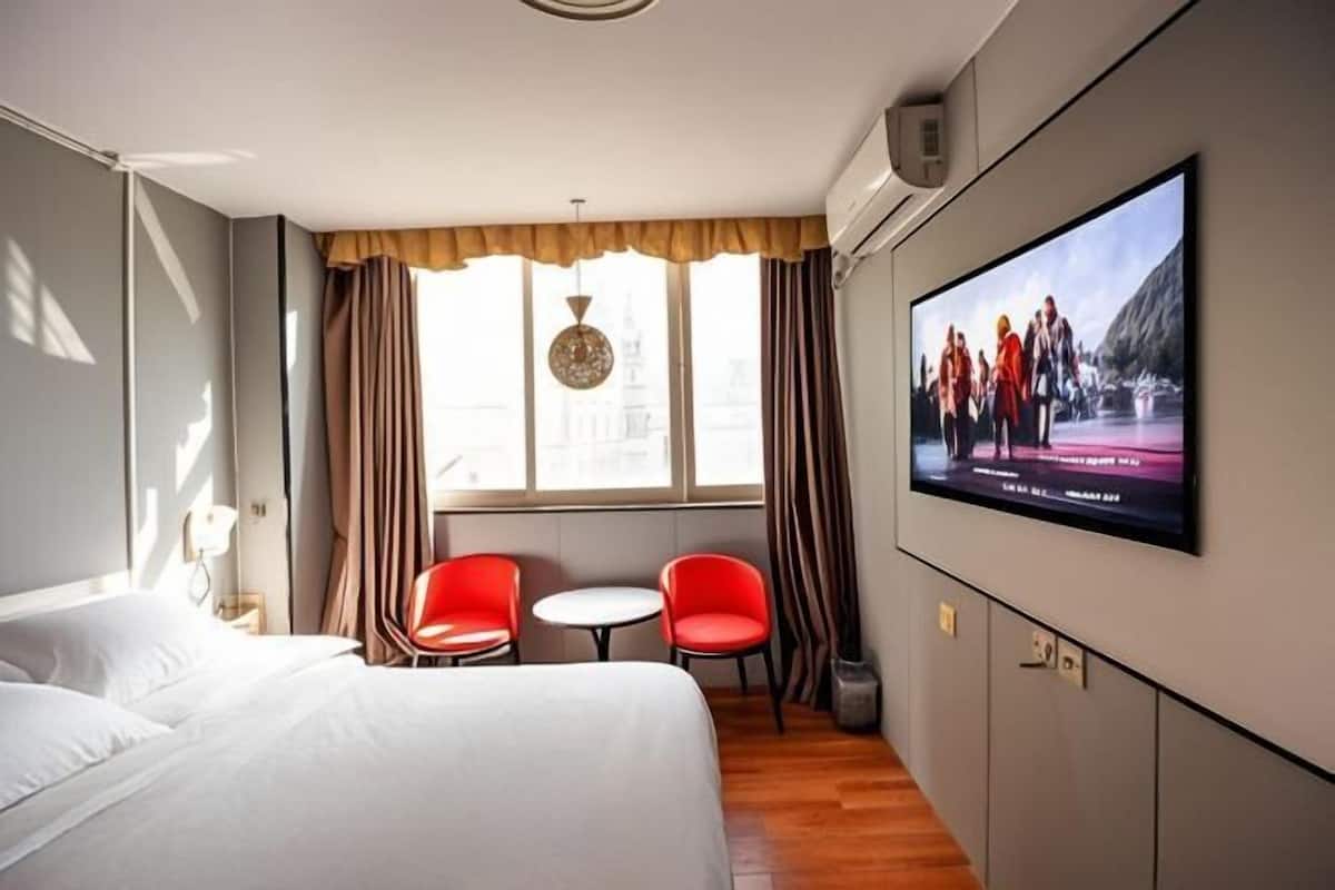 Habitación de hotel con una cama, dos sillas rojas, una mesita, un televisor en la pared y una ventana con cortinas.
