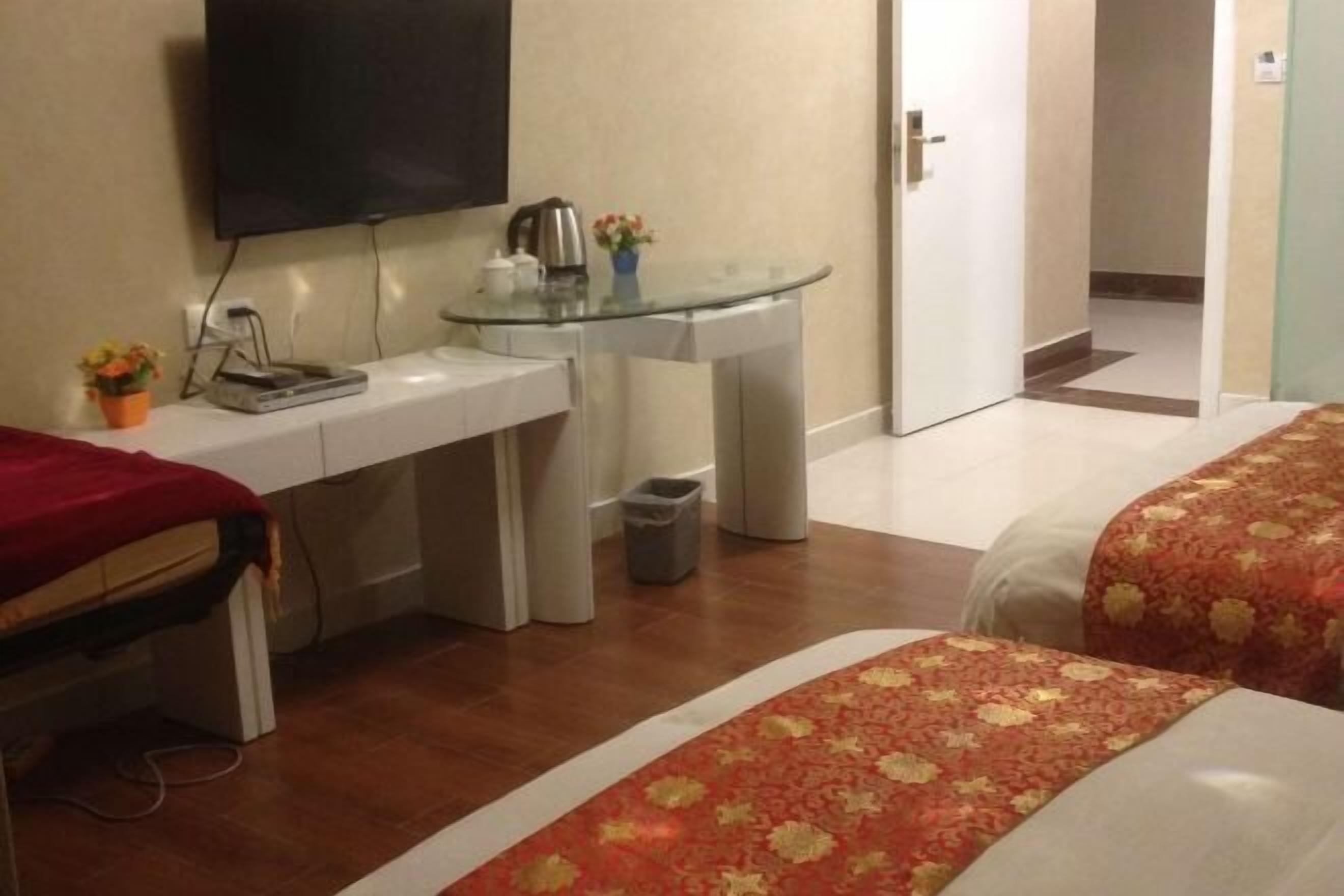 A hotel room with a bed, a desk with a television, a glass table, and a small trash bin.