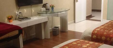 A hotel room with a bed, a desk with a television, a glass table, and a small trash bin.