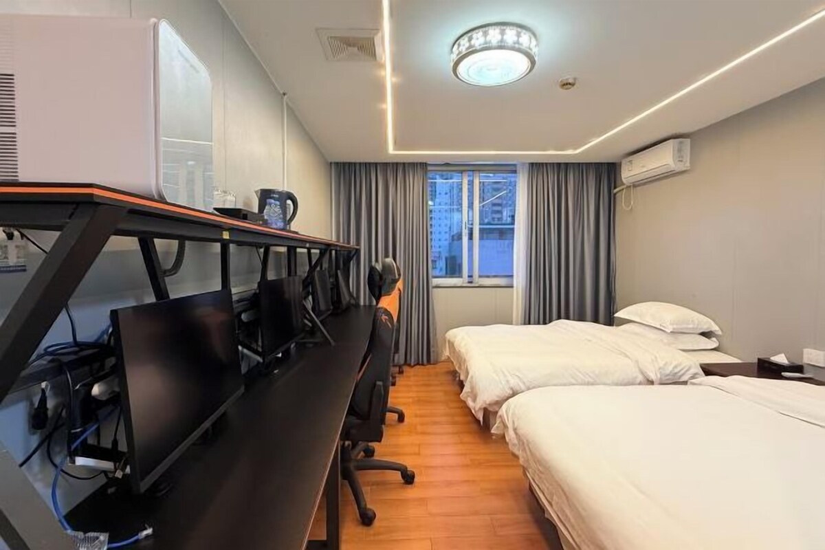 A hotel room with a desk, two beds, a window with curtains, and an air conditioning unit.