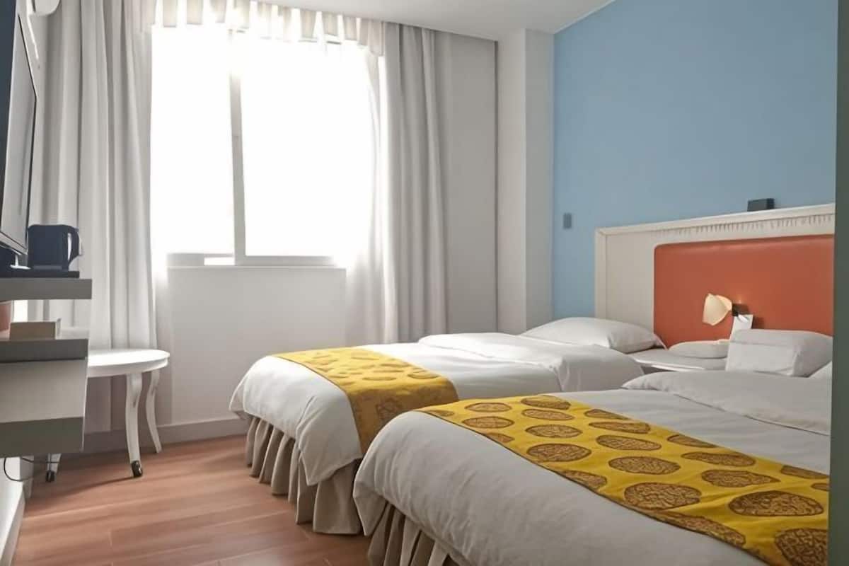 Una habitación de hotel con dos camas, un escritorio, una silla, un ventanal con cortinas y una mesita de noche con lámpara.