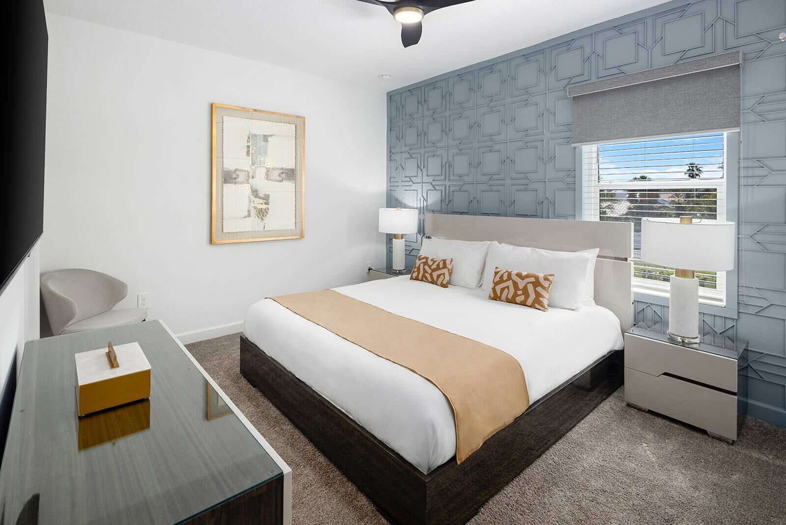 Habitación de hotel con cama, mesitas de noche, escritorio, silla, ventilador de techo, ventana con vistas y pared con diseño geométrico.