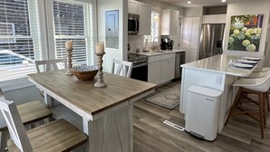 Une cuisine moderne avec des meubles blancs, un îlot central en bois doté de sièges et une grande fenêtre avec des stores.