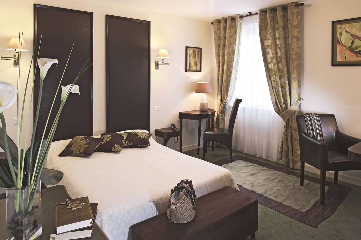 A hotel room with a bed, bedside table, lamp, chair, and a vase with flowers.