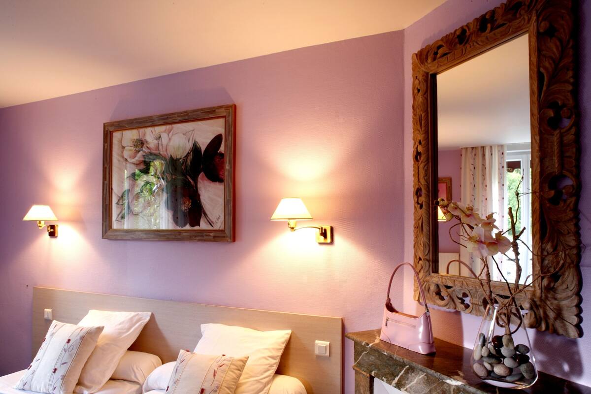 A hotel room with a bed, wall-mounted lamps, a painting, a mirror, and a vase with flowers.