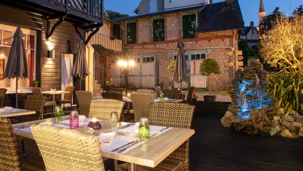 An outdoor dining area with wicker furniture, tables set with tableware, and a decorative water feature. - Hotel Le Normandie Spa (Bagnoles-de-l'Orne)