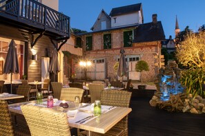 Un área de comedor al aire libre con mobiliario de mimbre, mesas dispuestas con vajilla y una fuente decorativa.