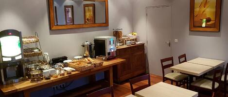 Un área de desayuno con una barra libre, que incluye una máquina de café, cestas de pan y una cafetera. Hay mesas y sillas de madera dispuestas para comer.