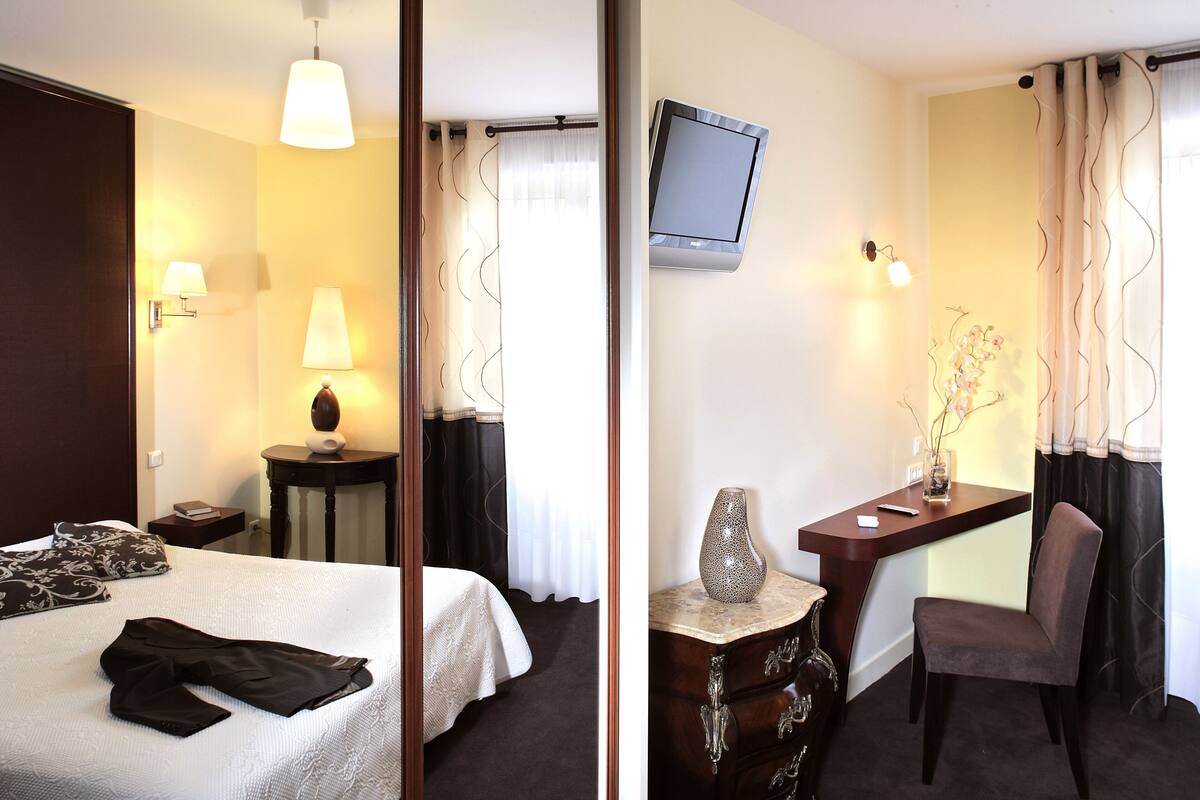 A hotel room with a bed, bedside table, lamp, TV, desk, chair, and a vase with flowers.