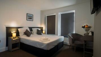 Quarto de hotel moderno com uma cama grande, luminárias nas laterais, uma mesa de trabalho com cadeira e uma janela com vista.