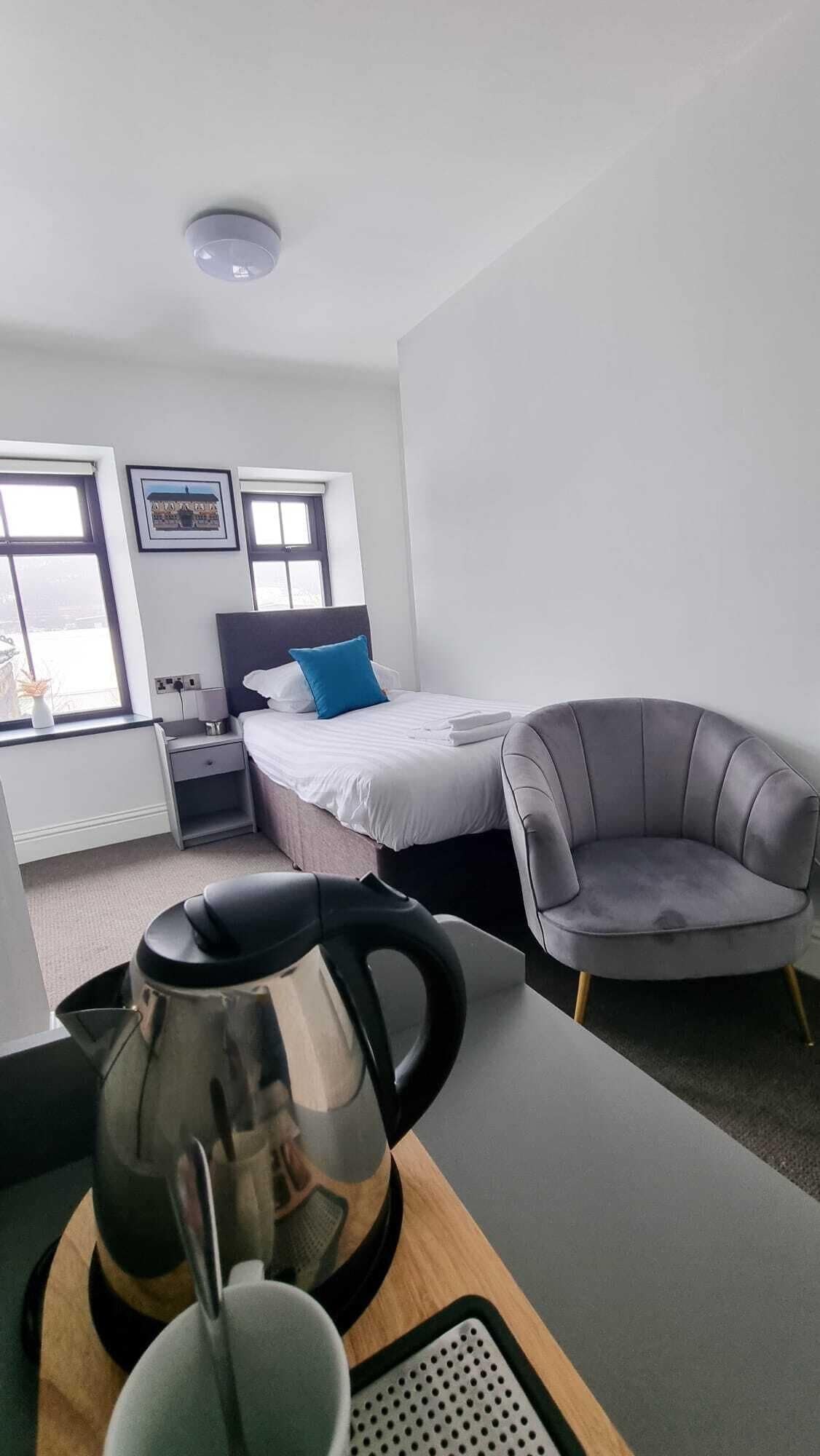 A modern hotel room with a bed, a gray armchair, a coffee table with a kettle, and a window with a view.