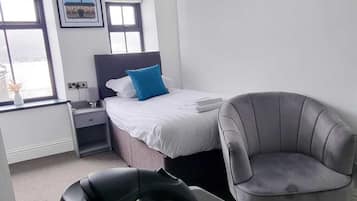 A modern hotel room with a bed, a gray armchair, a coffee table with a kettle, and a window with a view.