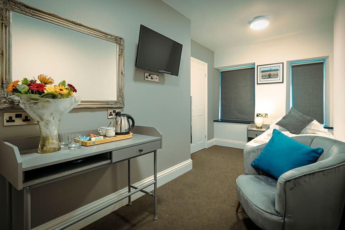 A hotel room with a desk, a chair, a bed with a blue pillow, a TV, and a window with blinds.