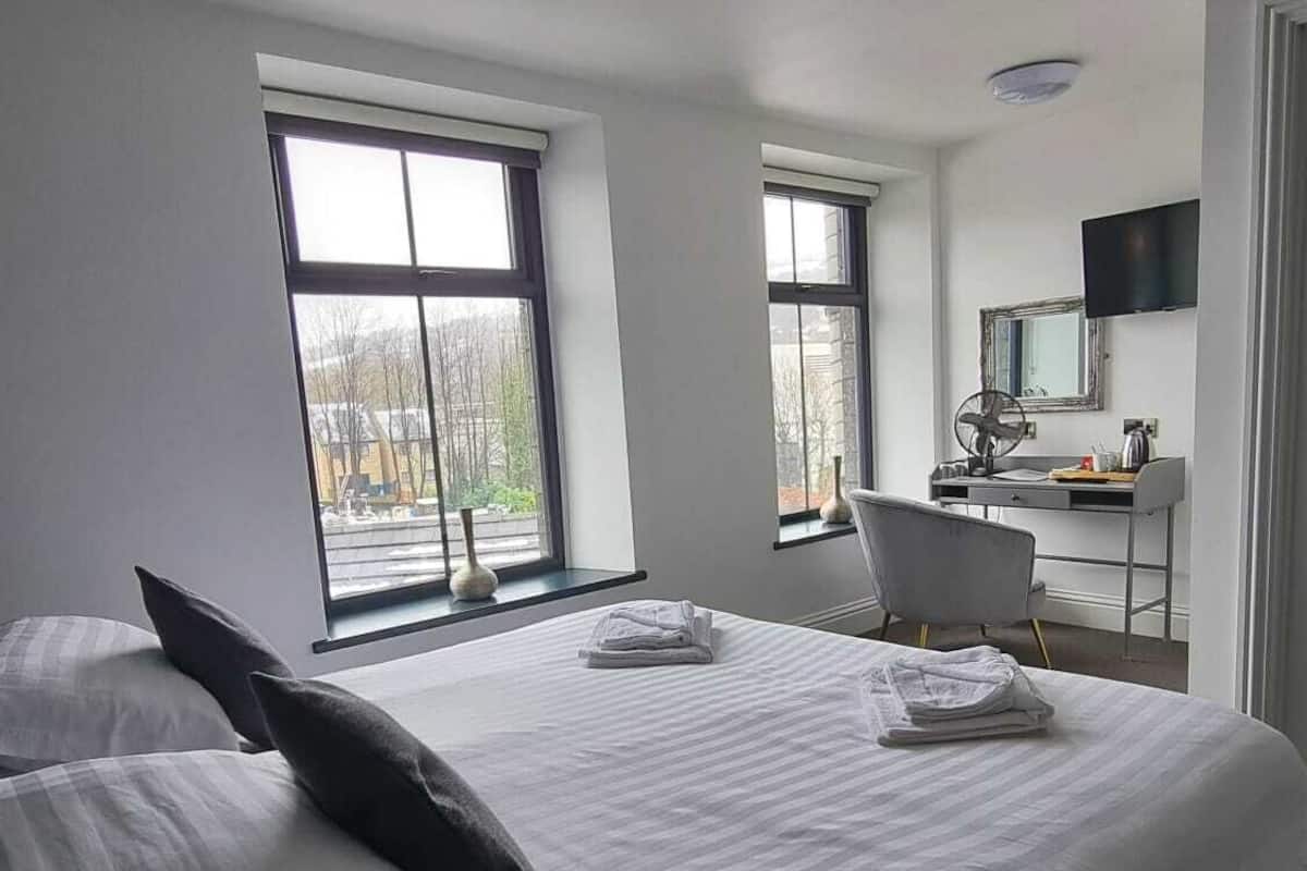 A modern hotel room with a large bed, two windows, a desk, and a television.