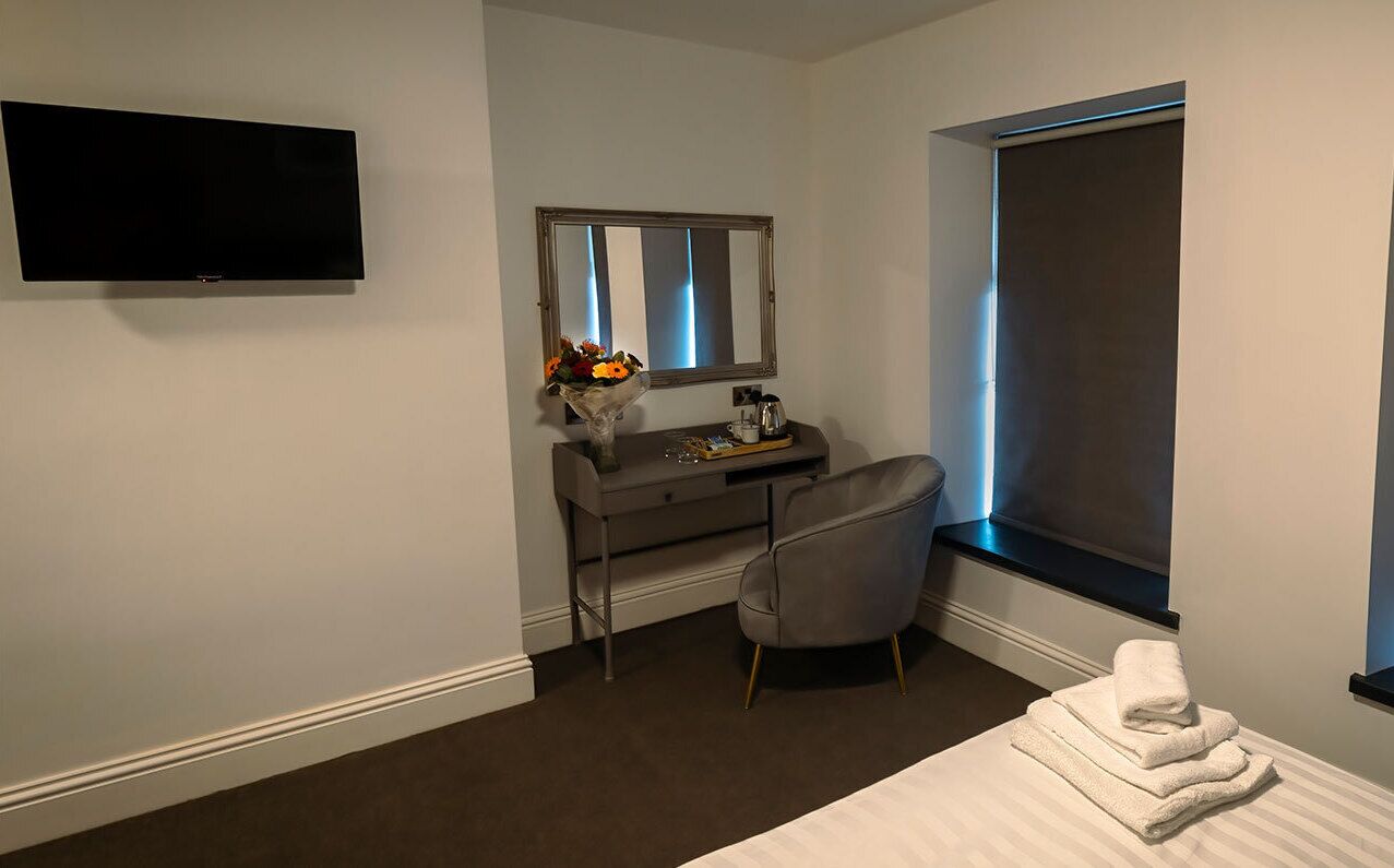 A hotel room with a flat-screen TV mounted on the wall, a vanity with a mirror, a chair, and a window with blinds.