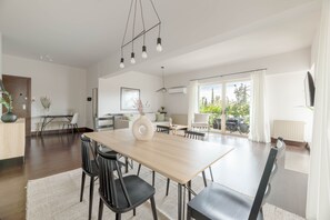 A modern dining area with a wooden table and black chairs, a kitchen in the background, and a view of greenery through large windows. - Panoramic 1BR Apt near Kallimarmaro (Athens)