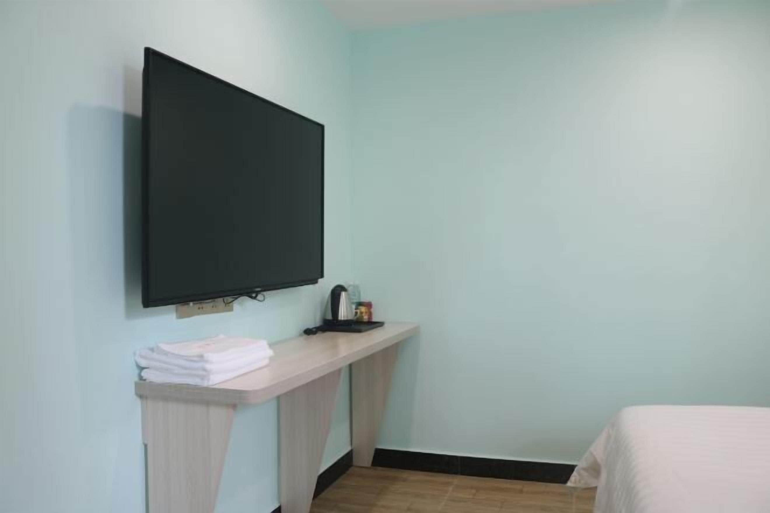 A room with a bed, a TV mounted on the wall, a small table with a kettle, and a stack of white towels.