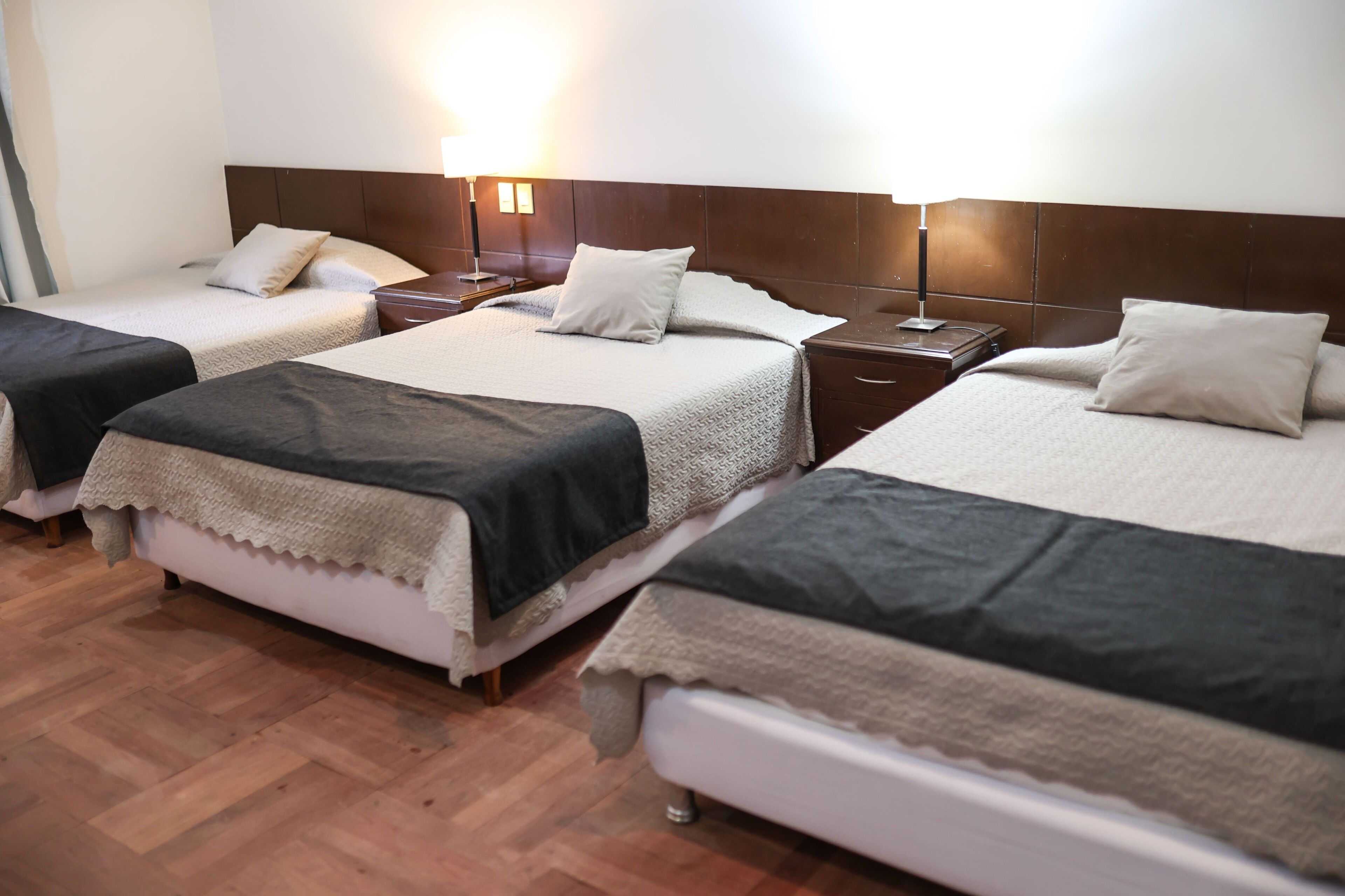 Une chambre d’hôtel avec trois lits simples, un plancher en bois et des tables de chevet avec des lampes.