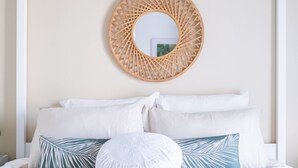 A neatly made bed with white and blue patterned pillows, a round woven mirror on the wall, and a rolled beige towel on the bed.
