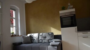 A living room with a grey sofa, a small table with a plant, a window with red flowers, and a kitchen area with a microwave and oven. - Apartment "Boskoop" with terrace - Apartments Liebisch (Flieth-Stegelitz)