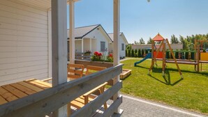 Un espace extérieur aménagé, abrité et doté de bancs en bois et d’une table, une aire de jeux avec un toboggan et des balançoires, et un bâtiment à l’arrière-plan, avec une façade blanche et un toit rouge.