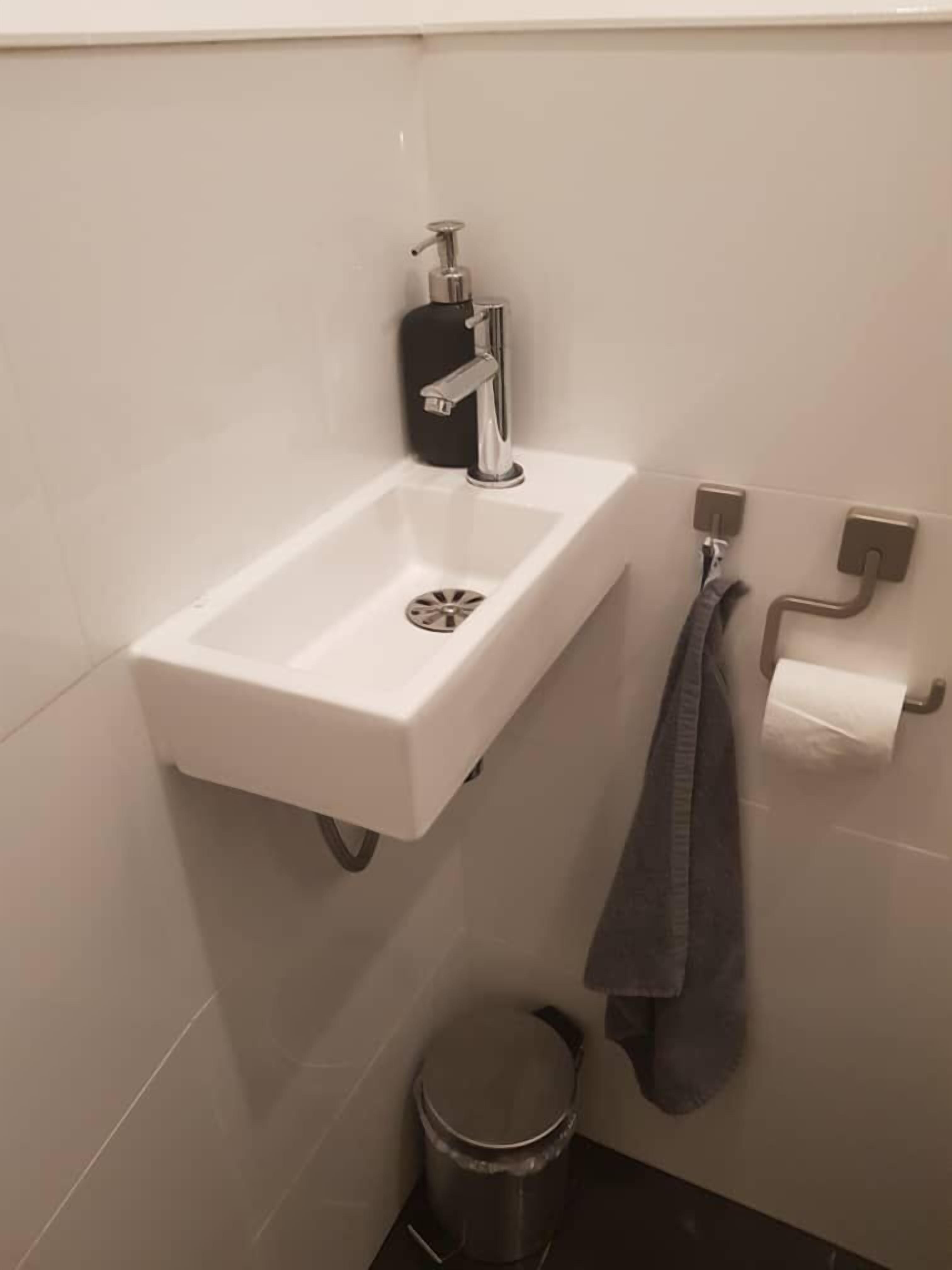 Een badkamer met een witte wastafel, een zwarte zeepdispenser, een handdoekhouder met een handdoek en een prullenbak.