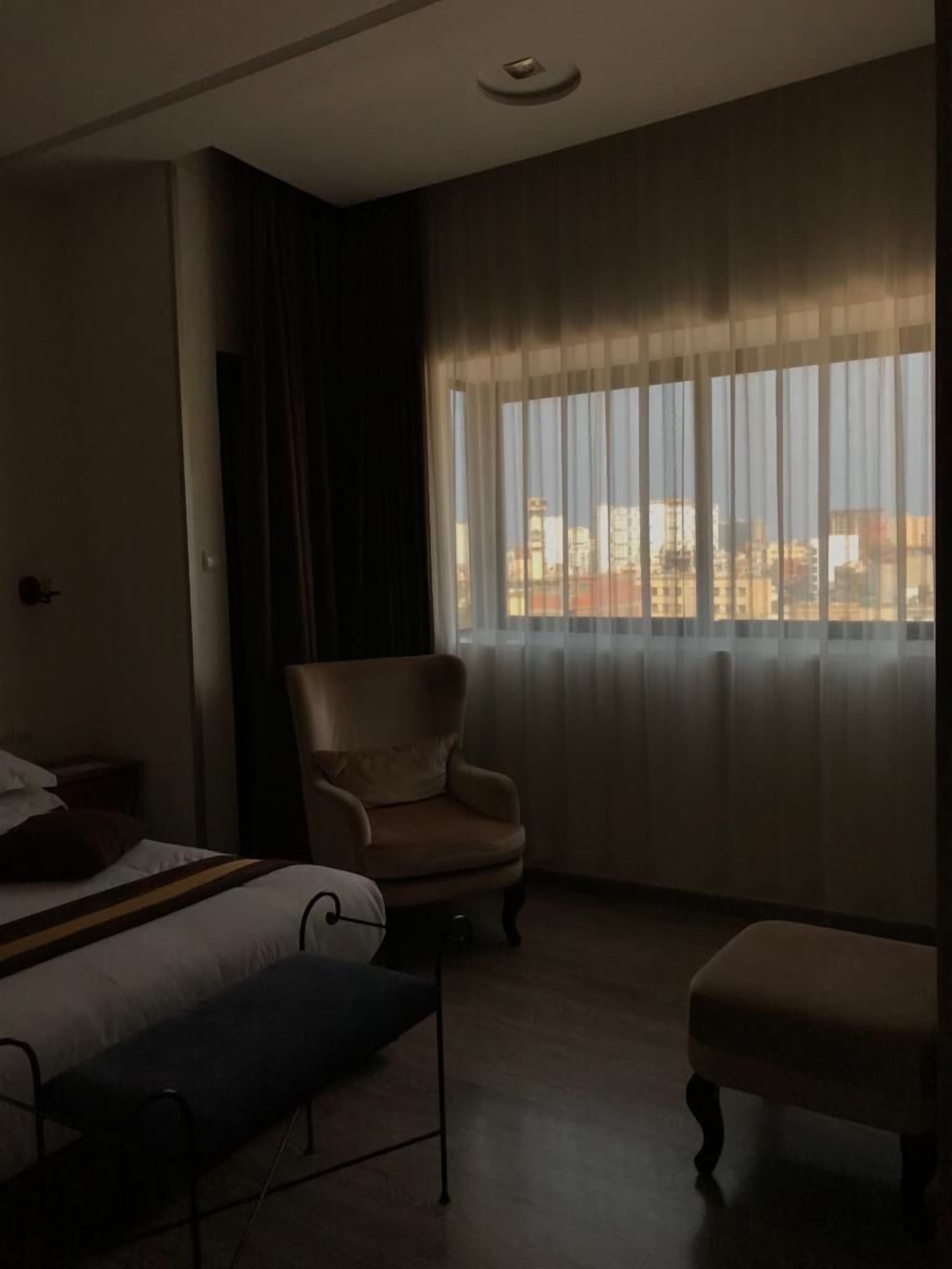 Una habitación de hotel con una cama, una silla, un reposapiés y un ventanal con cortinas.