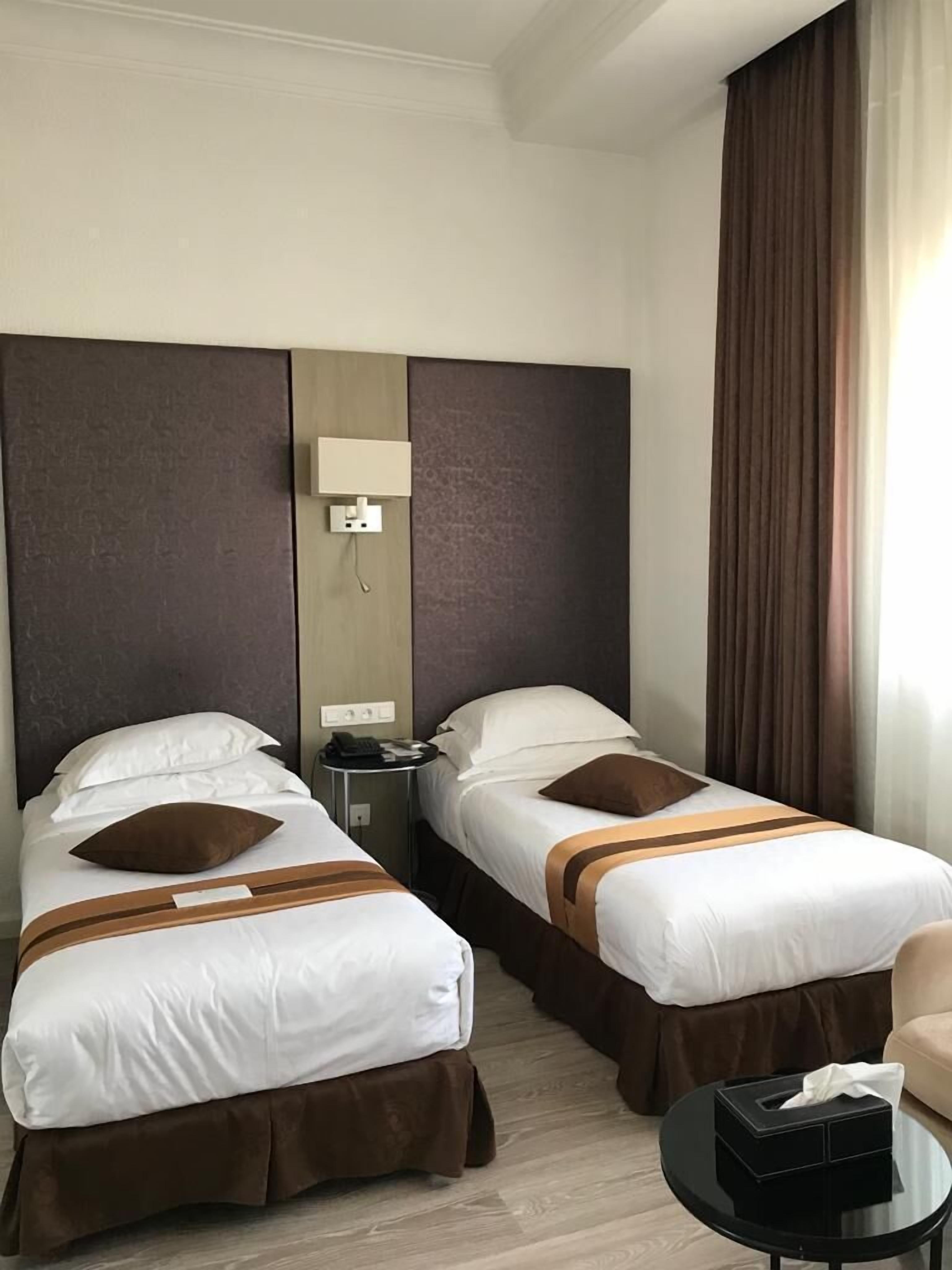 A hotel room with two single beds, a bedside table with a lamp, a small round table with a tissue box, and a large window with curtains.