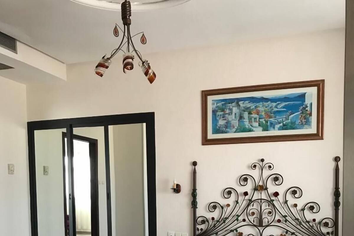 A bedroom with a bed, a bedside table, a nightstand, a painting, and a chandelier.
