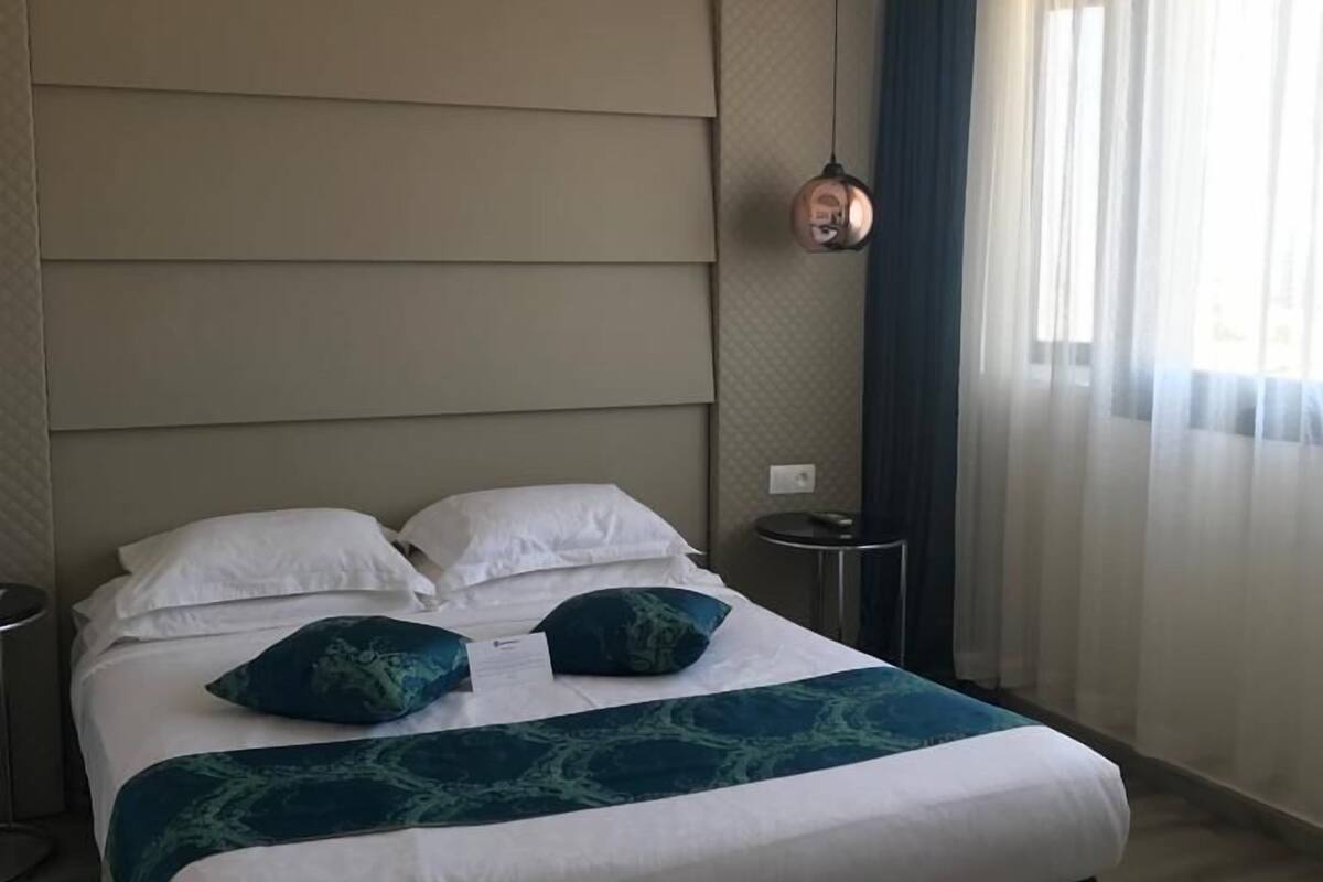 A modern hotel room with a large bed, a headboard with horizontal panels, a round bedside table, and a hanging pendant light.