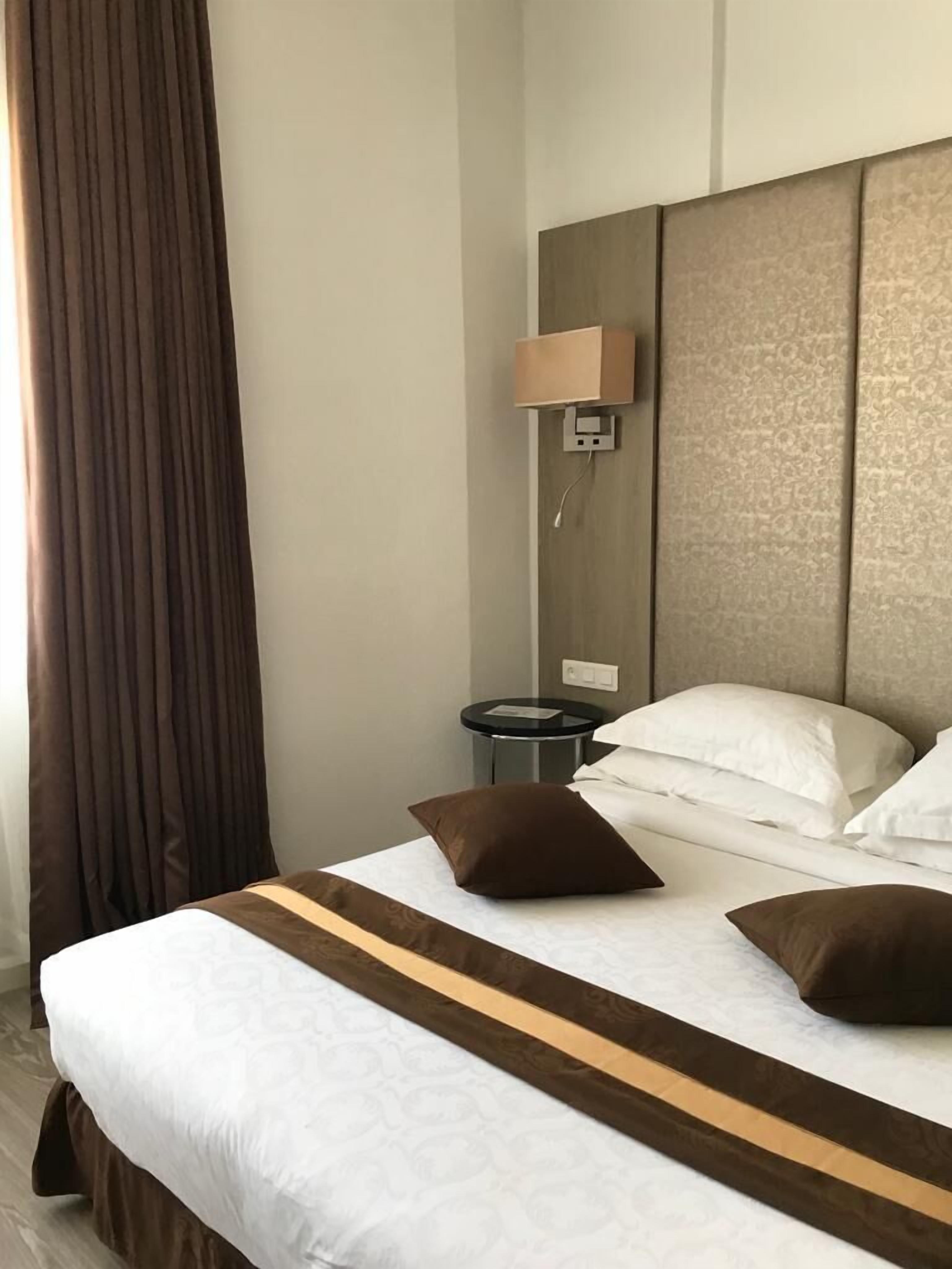 Habitación de hotel con una cama grande, una mesita de noche, una lámpara de pared y vistas al exterior a través de una ventana con cortinas.