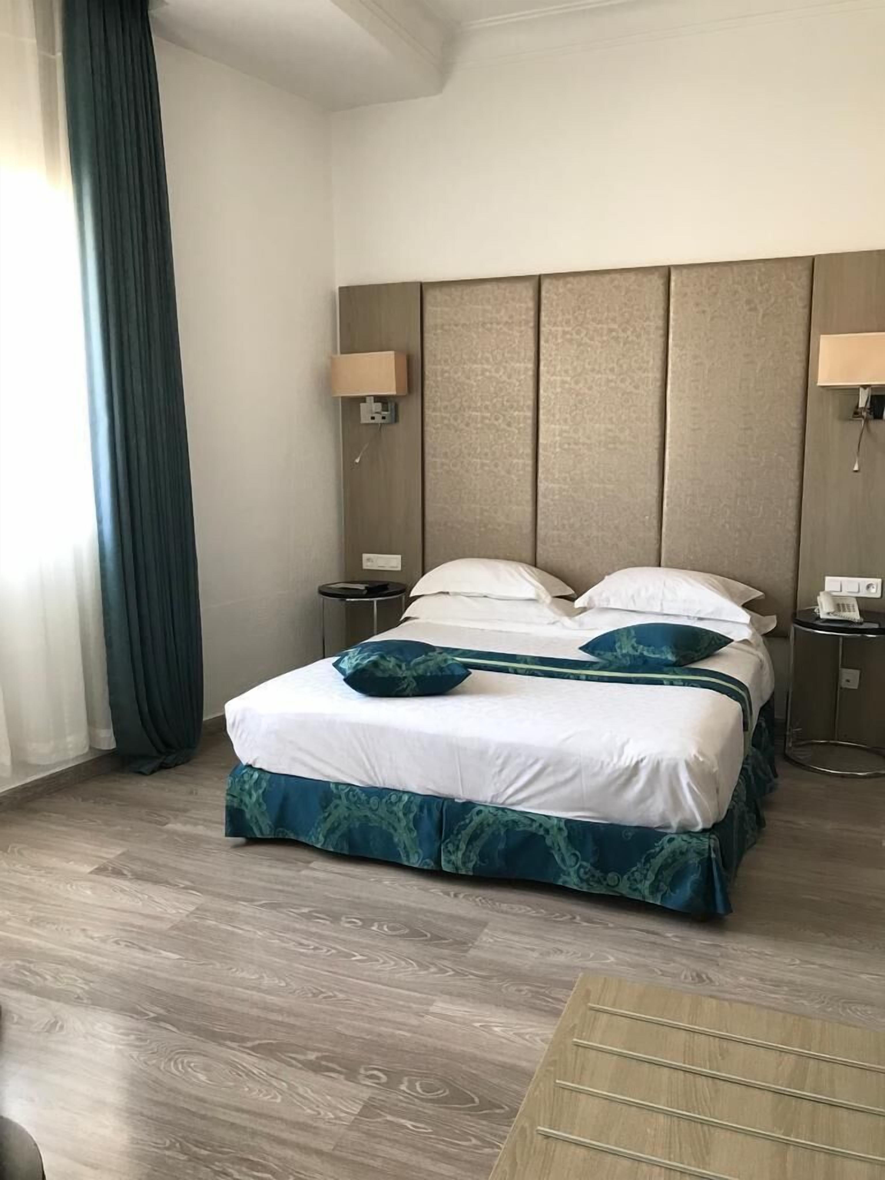 A hotel room with a large bed, two bedside tables, a nightstand, a wooden floor, and a window with curtains.