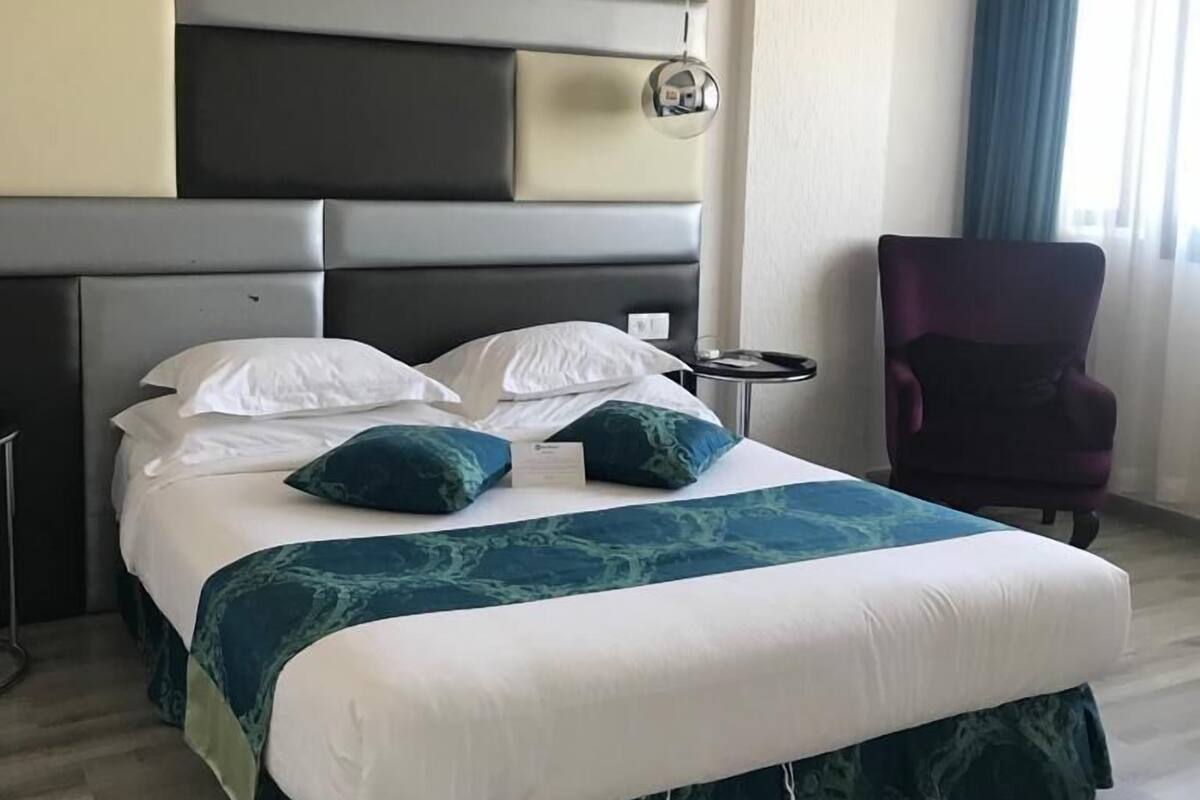 A modern hotel room with a large bed, a headboard with storage compartments, a small bedside table, and a chair.