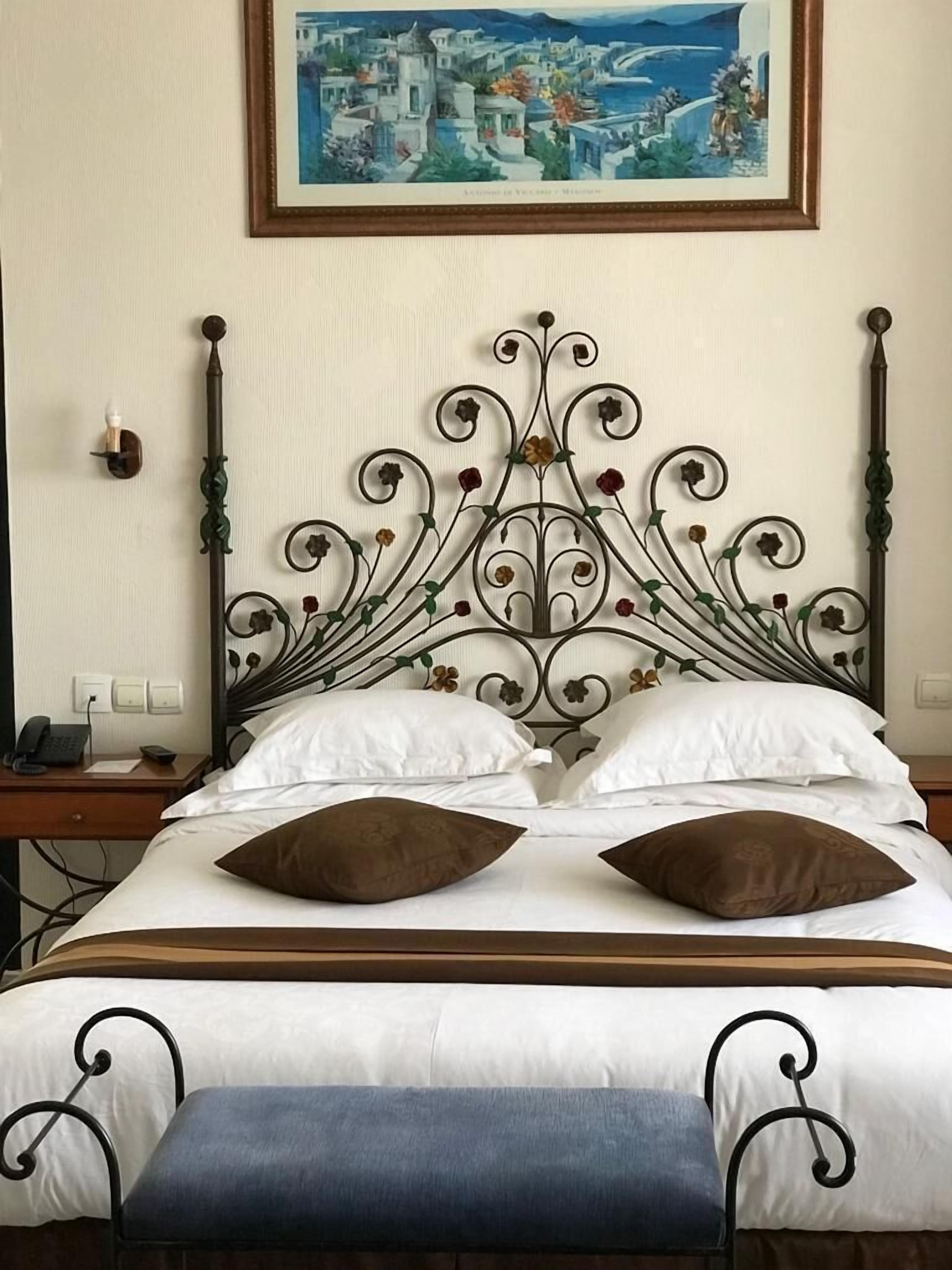 A neatly made bed with a decorative metal headboard, white pillows, and a brown cushion. A painting of a coastal town hangs above the bed.