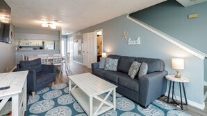 A living room with a gray sofa, a coffee table, and a patterned rug. There is a kitchen in the background with white cabinets and a microwave. - Coastal Condo | Beach Views, Hot Tub & Gas Grills (Corpus Christi)