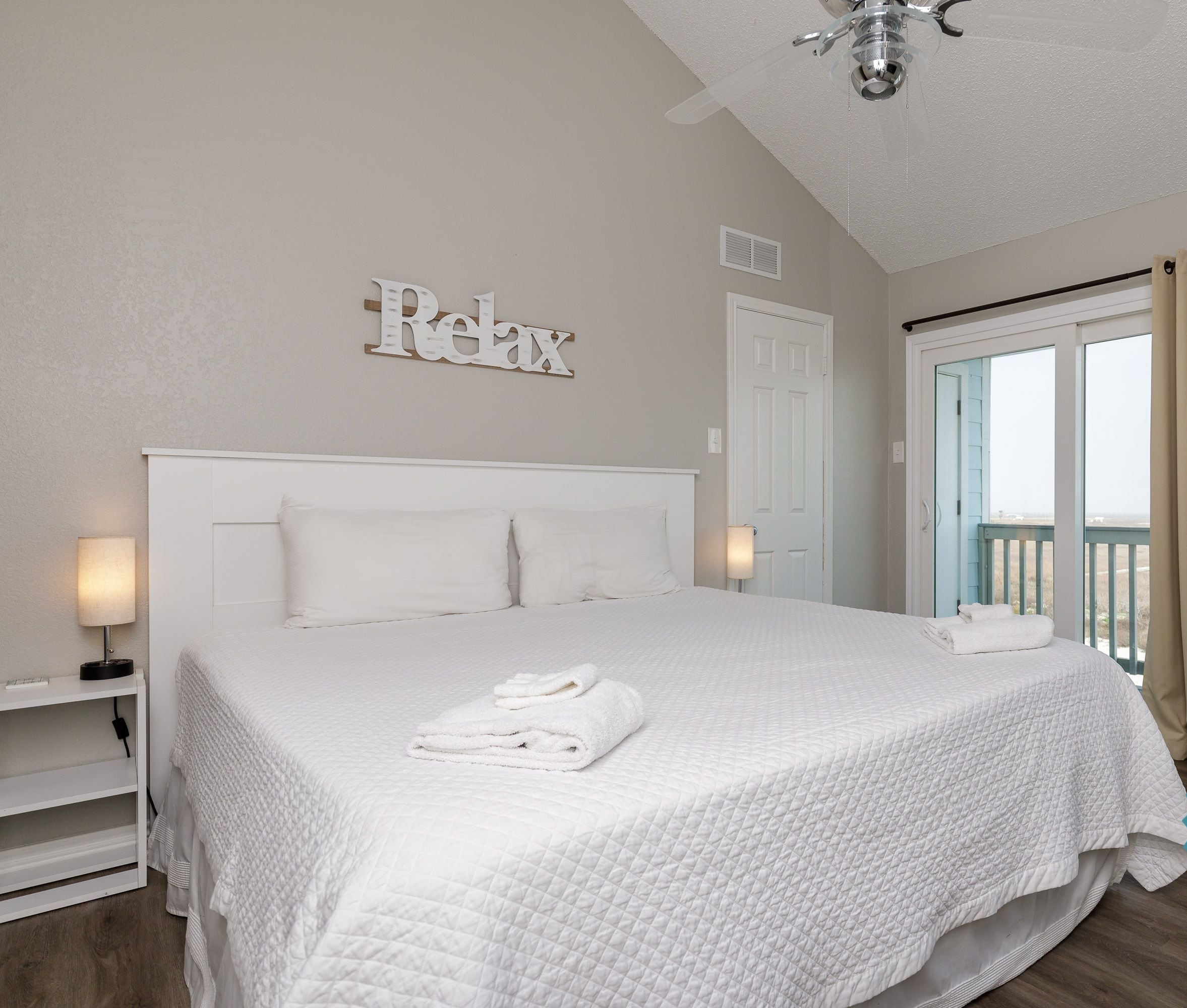 A clean, white-themed bedroom with a bed, a ceiling fan, and a view of the outdoors.