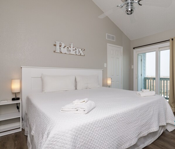 A clean, white-themed bedroom with a bed, a ceiling fan, and a view of the outdoors.
