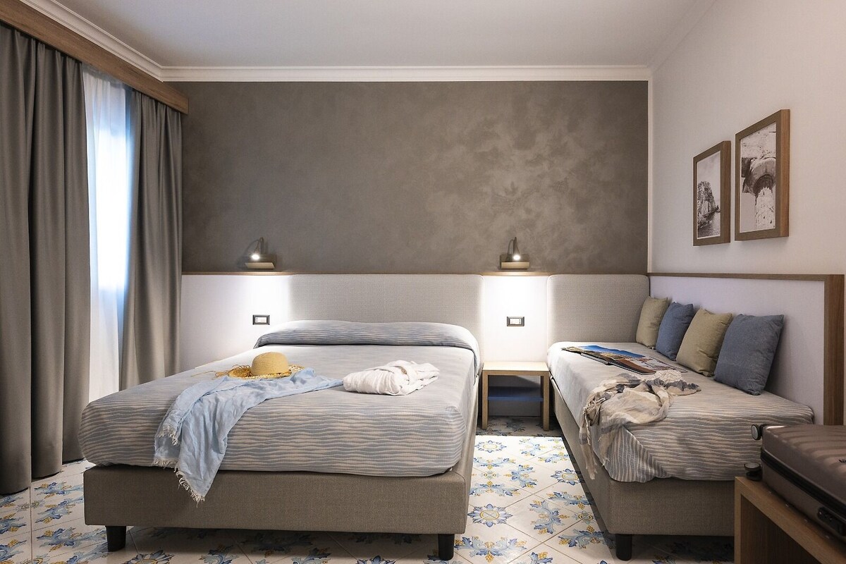 A hotel room with two beds, a small table, and a bench. There are two bedside lamps and a wall-mounted shelf with a lamp. The room has a large window with curtains and a tiled floor.