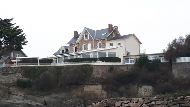 A large, multi-story house with a stone facade and a sloped roof, situated on a rocky embankment by the sea.