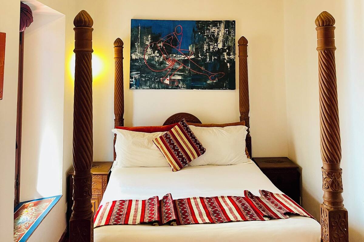 A bedroom with a four-poster bed, a chandelier, and a cityscape painting on the wall.