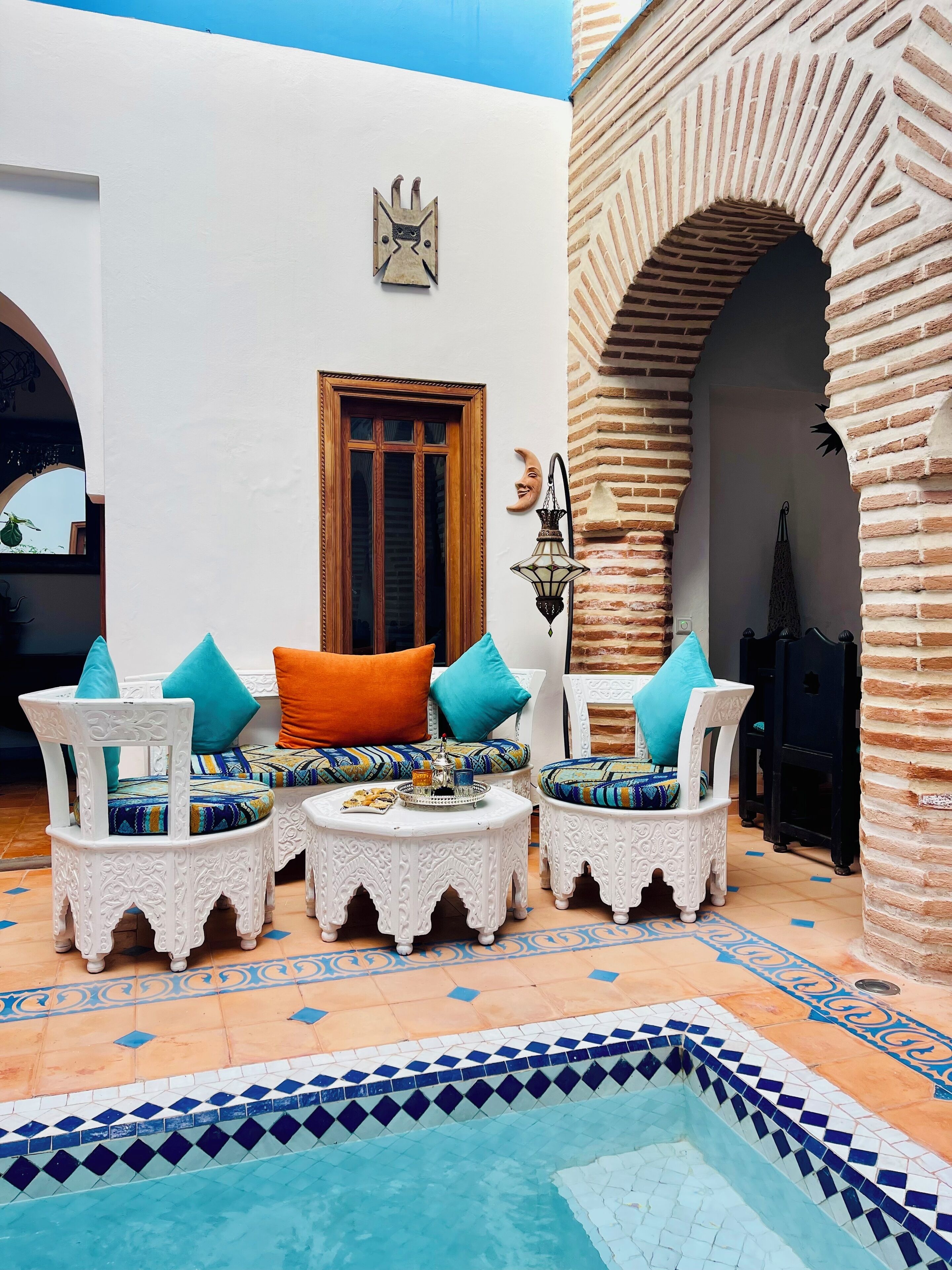 A cozy outdoor seating area with white ornate chairs and cushions, a small pool with blue mosaic tiles, and a brick archway.