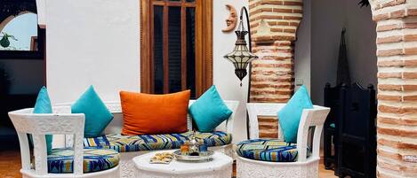 A cozy outdoor seating area with white ornate chairs and cushions, a small pool with blue mosaic tiles, and a brick archway.