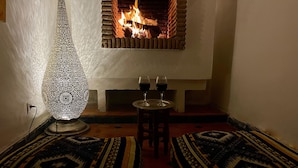 A cozy room with a fireplace, two patterned rugs, a tall decorative lamp, and two glasses of red wine on a small table. - Riad Zarka (Marrakech)