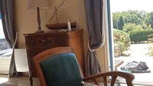 Une pièce avec une chaise en bois, une commode, un tableau et une vue sur la verdure à l’extérieur.