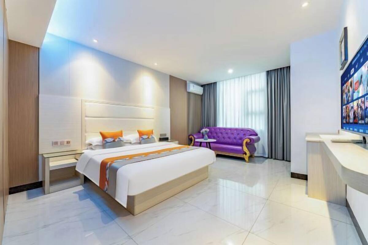 A hotel room with a large bed, a purple sofa, a desk, and a TV.