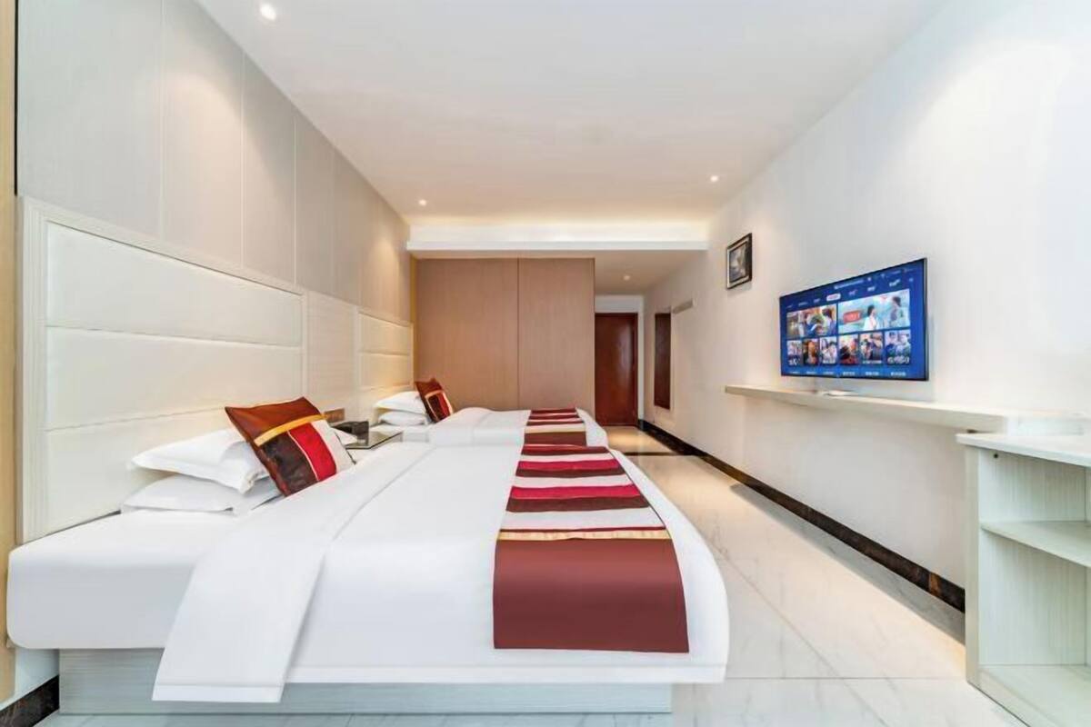 A hotel room with a long bed, a television on the wall, and a hallway leading to another room.