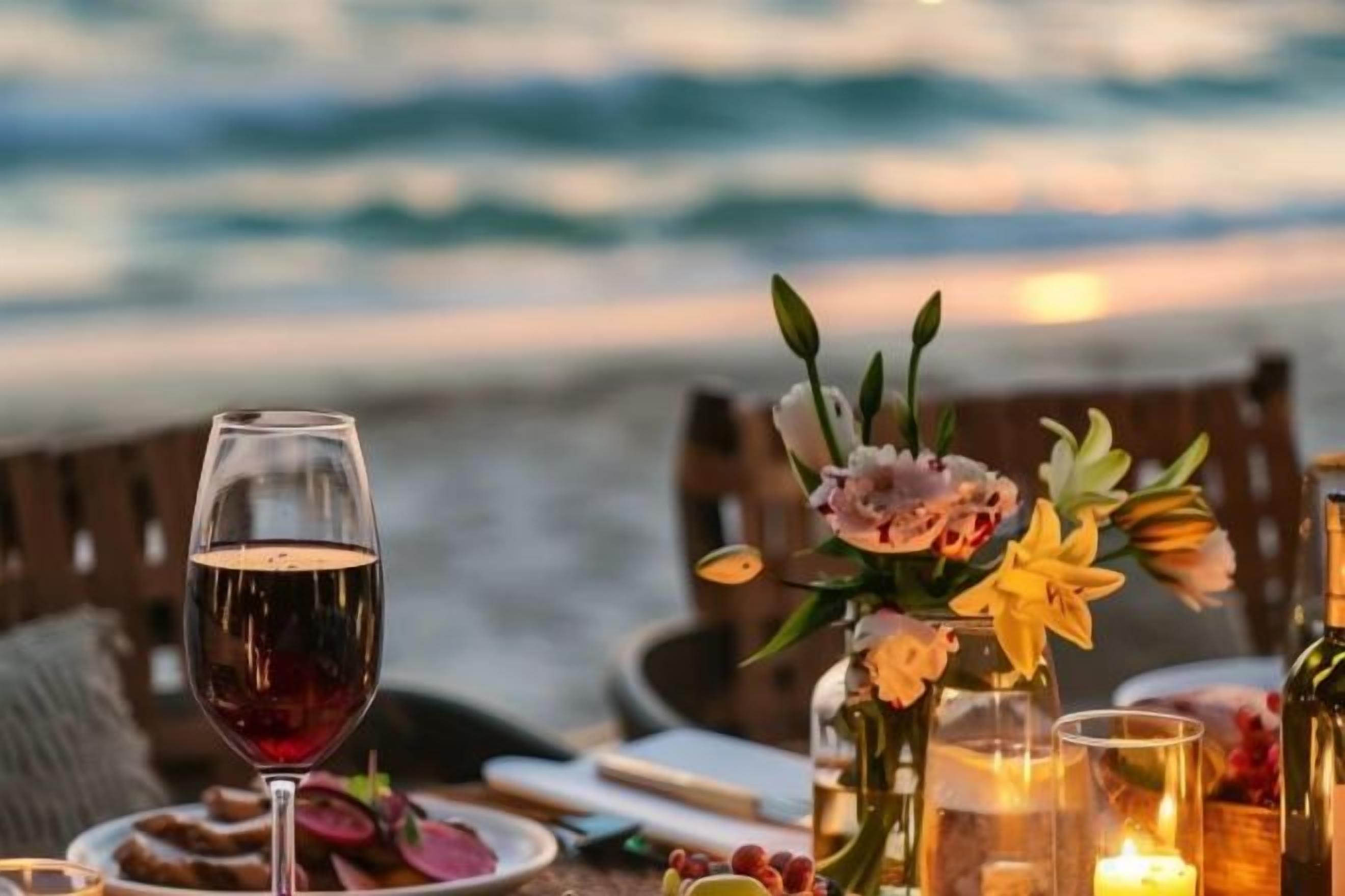 Een gedekte tafel met een glas rode wijn, kaarsen en bloemen, met uitzicht op een strand en golven.